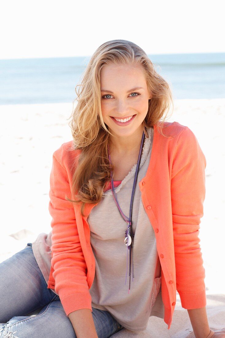 A young dark blonde woman on a beach wearing a top, a cardigan and jeans