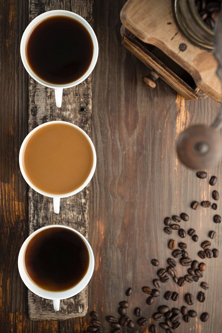 An arrangement of coffee featuring three cups of coffee, coffee beans and a coffee grinder