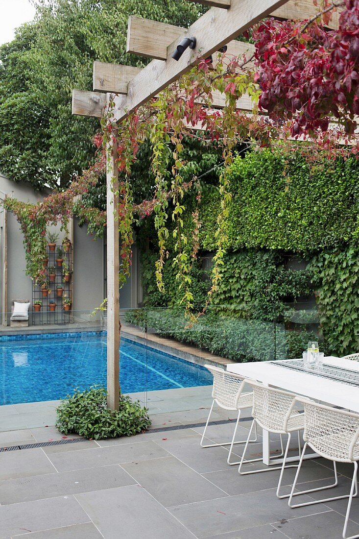 Wooded pergola over white table and chairs in front of glass balustrade and pool