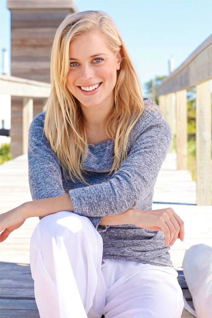 A young blonde woman on a wooden jetty wearing a grey jumper and white trousers