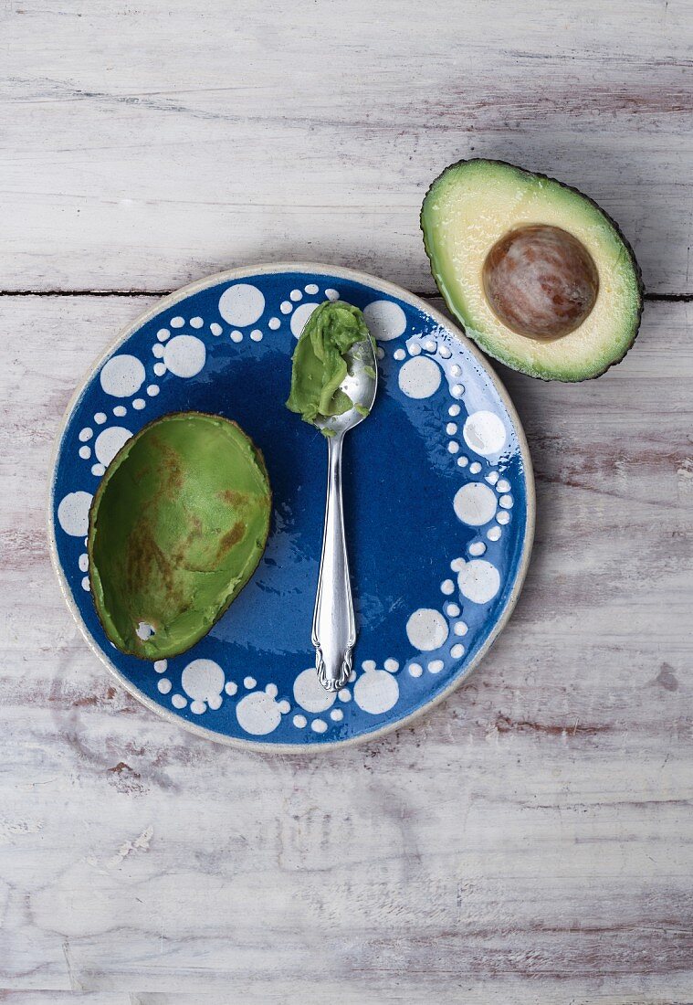 Halbe ausgeschälte Avocado auf blauem Tonteller mit Löffel und Resten von Avocado, daneben Avocadohälfte