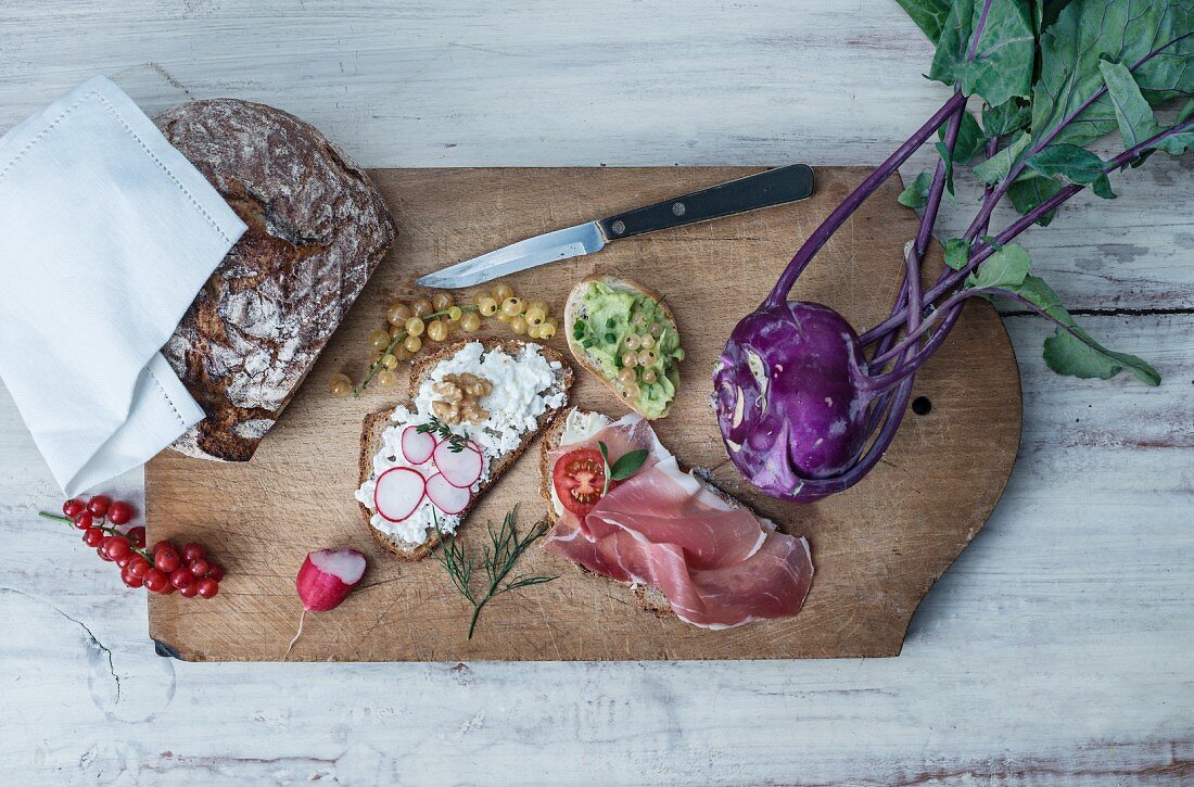 Slices of bread with various toppings on a chopping board with redcurrants, purple kohlrabi and crusty bread