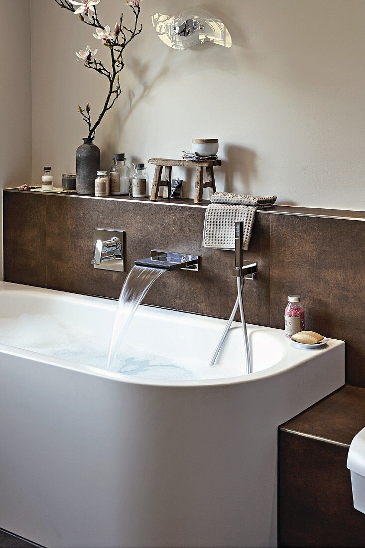 A designer bathtub with a fountain tap and shower head attached to a tiled wall panel