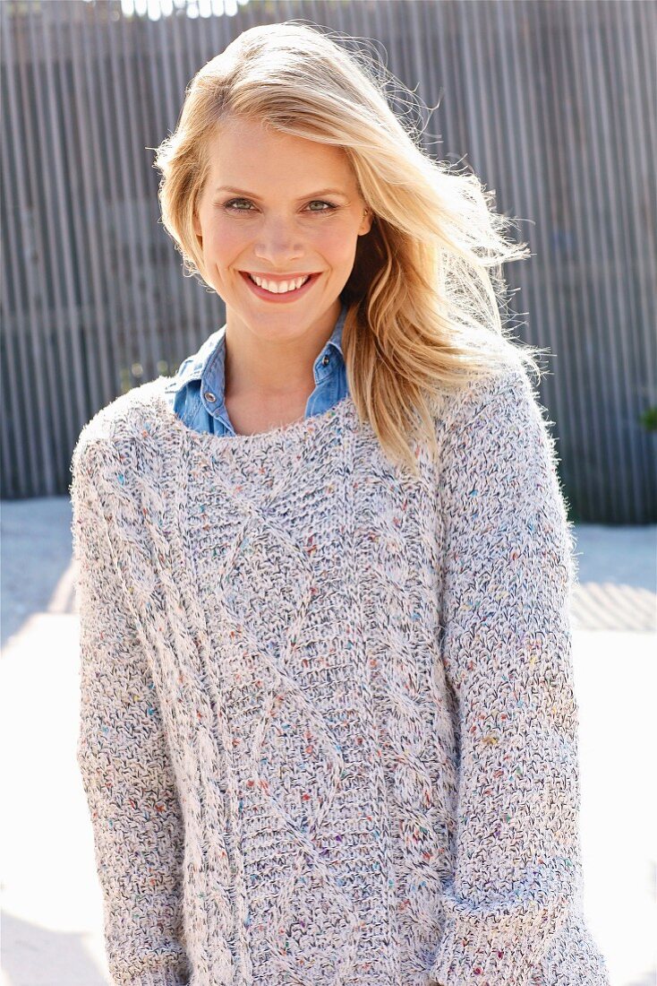 A blonde woman wearing a denim blouse with a coarse-knit jumper over her shoulders