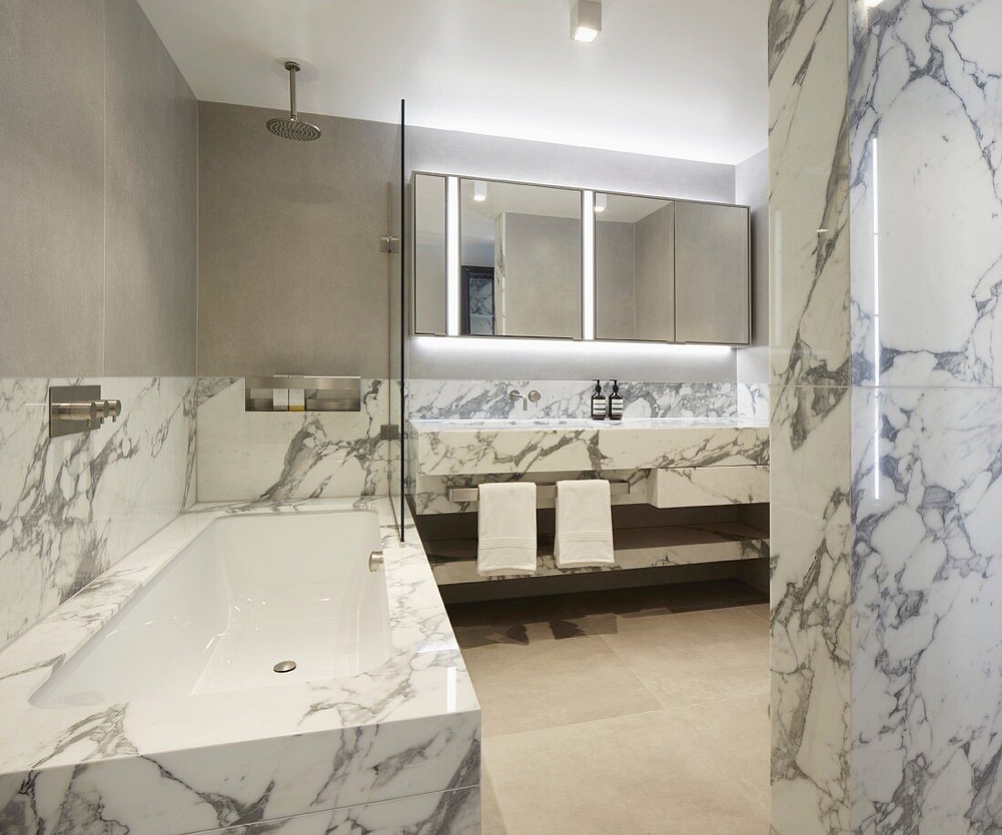 Marble-clad bathtub and washstand and backlit mirrored cabinet in luxurious bathroom