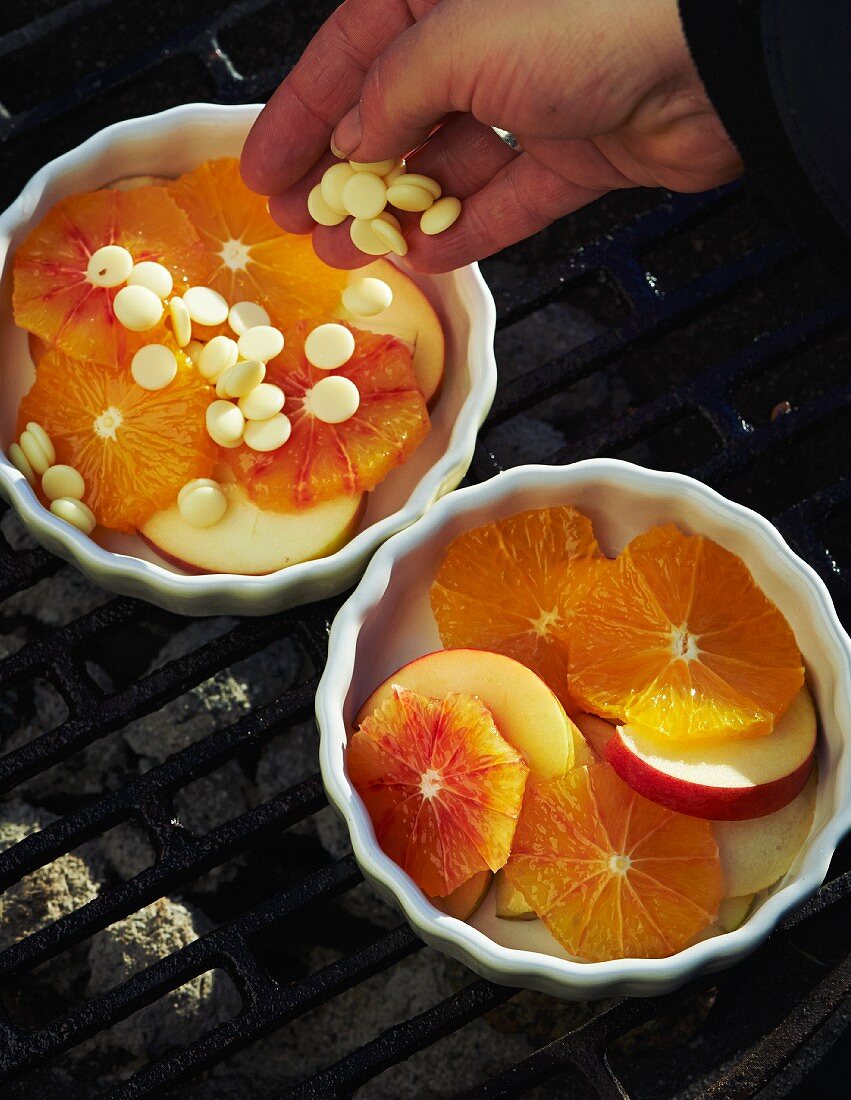 Hand gibt weiße Schoko-Chips über Fruchtscheiben in Gratinförmchen