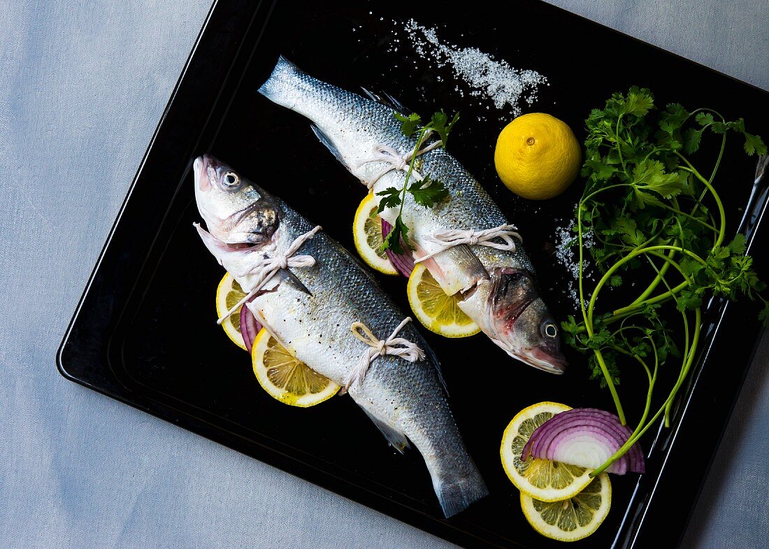 Two branzino fish stuffed with herbs, onion and lemon slices on a baking tray