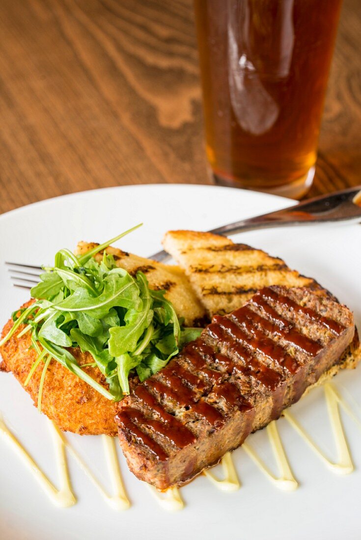 Grilled bread with meatloaf and rocket on a plate in front of the glass of beer