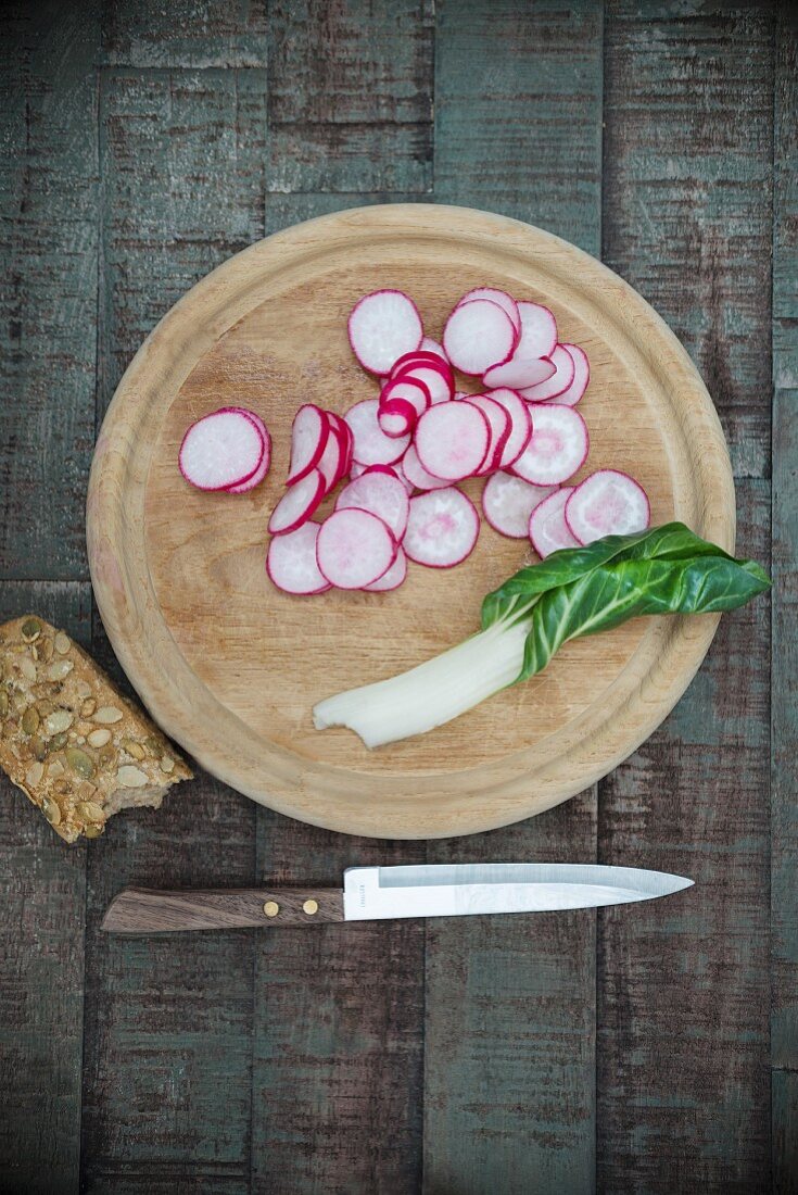 Sliced radishes and a chard leaf on a wooden board next to a knife and a sunflower seed roll