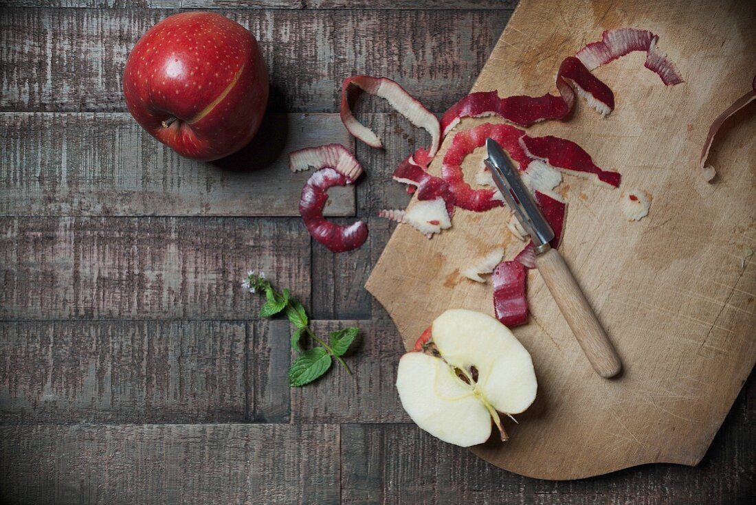Jonared apples with peel, an apple wedge, a peeler, fresh mint and a chopping board on a wooden surface