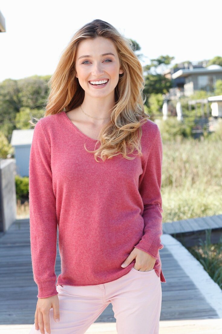 A young blonde woman wearing a pink knitted jumper and a pair of light trousers