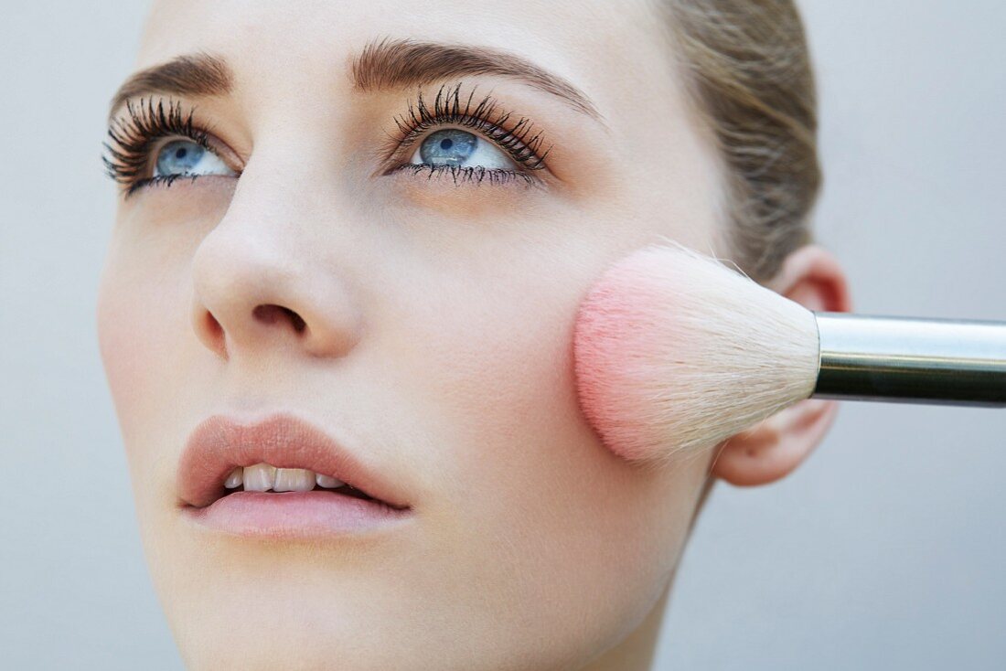 A young blonde woman holding a blushed brush against her cheek