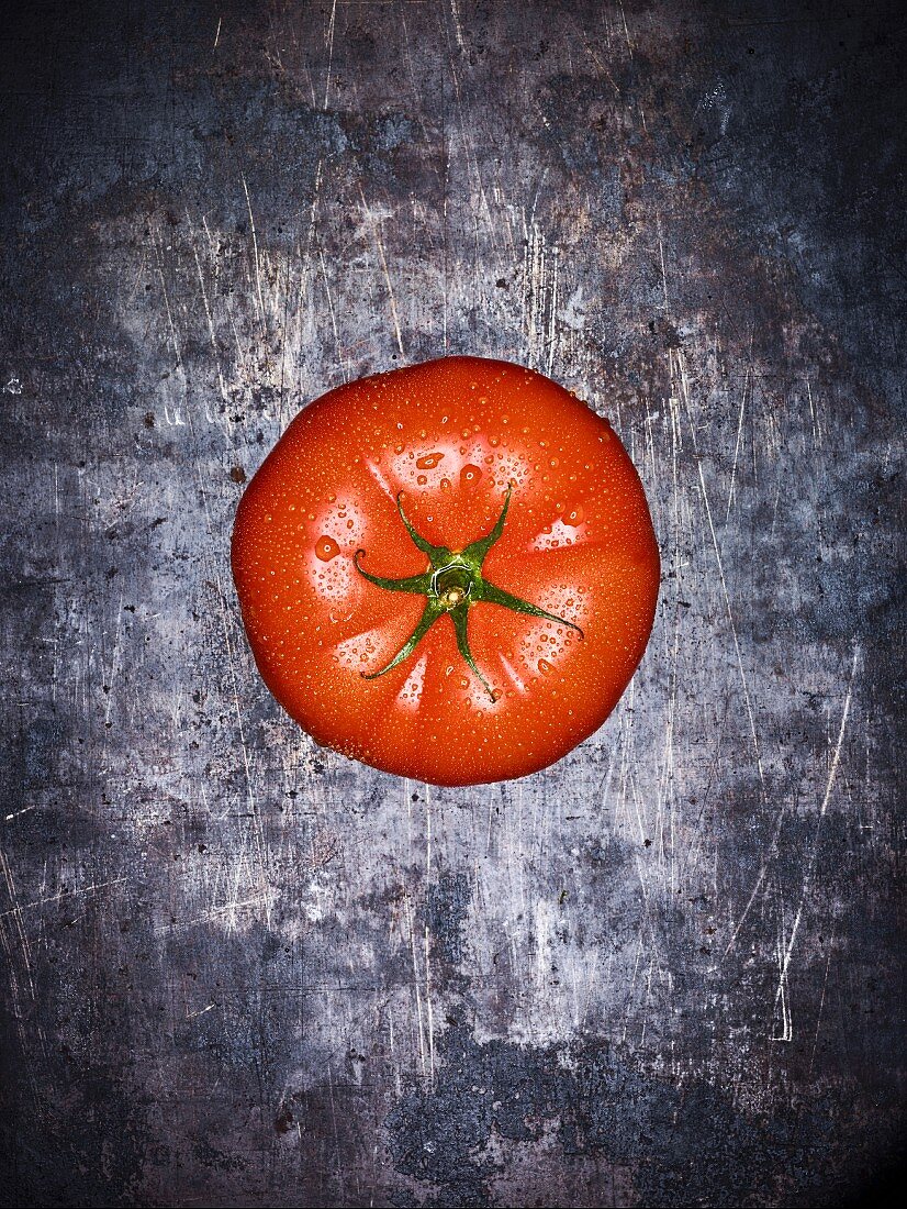 A beef tomato with droplets of water on a grey surface (seen from above)