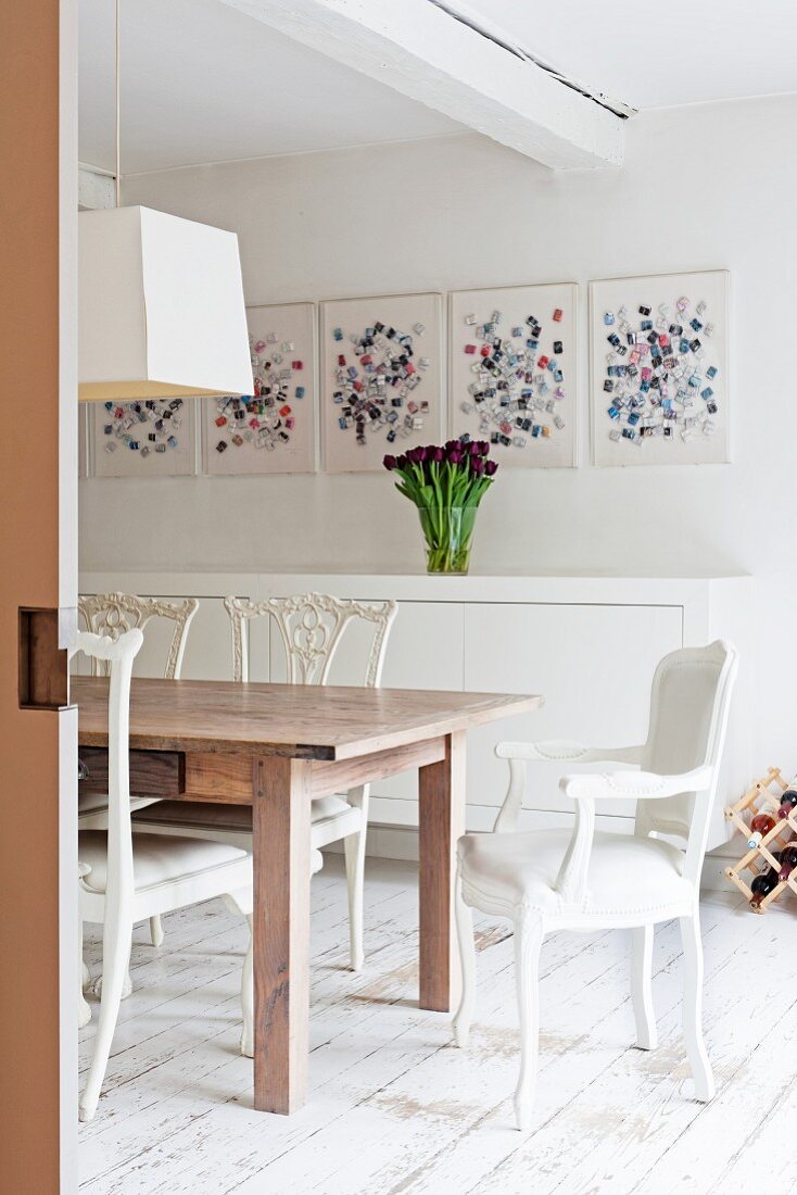 Wooden table, white chairs, white sideboard and modern art in dining area with vintage wooden floor