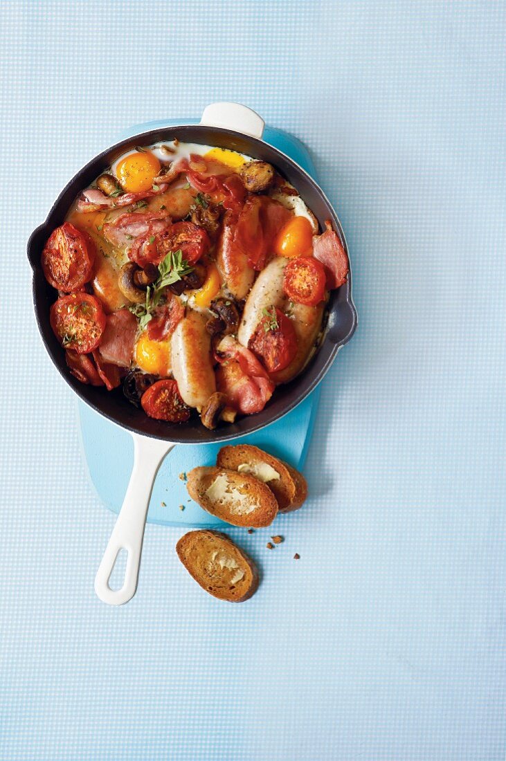 Reichhaltiges Frühstück mit Würstchen, Eiern, Speck, Tomaten und Champignons in einer Pfanne