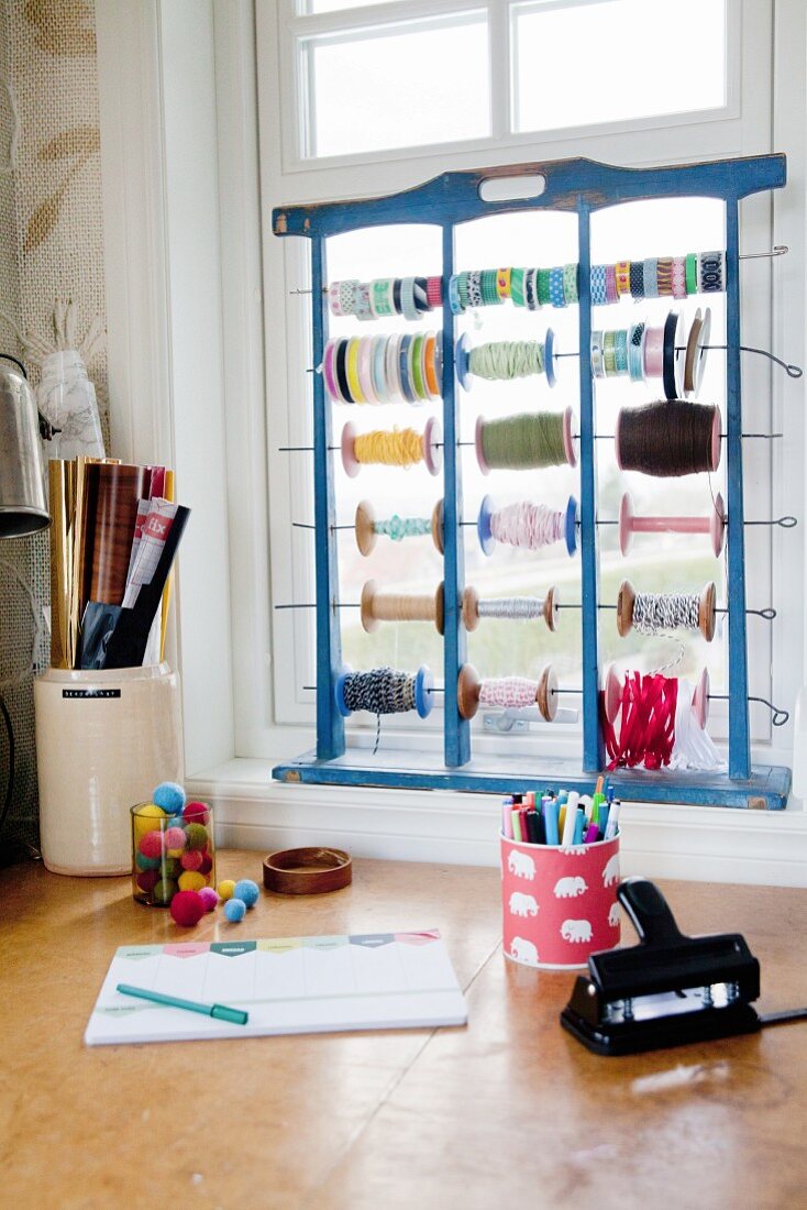 Craft materials and wooden frame holding reels of string and ribbon on windowsill above desk