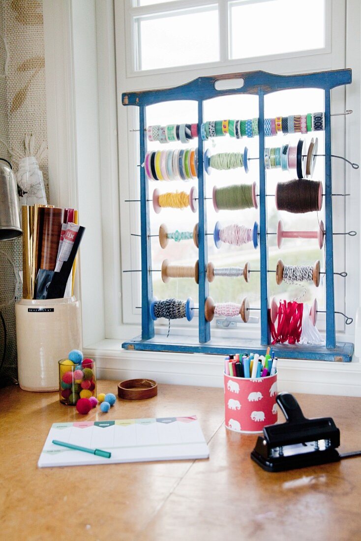 Schreibtisch mit Bastelmaterialien und Holzgestell mit bunten Garnrollen und Geschenkbändern auf Fensterbrett