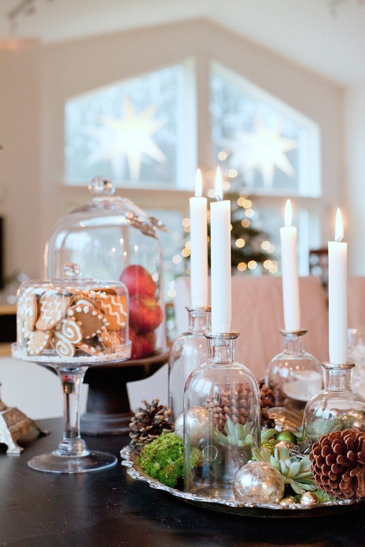 Selbst gestalteter Adventskranz mit vier brennenden Kerzen in Apothekerflaschen, Lebkuchen und Äpfel unter Glashauben