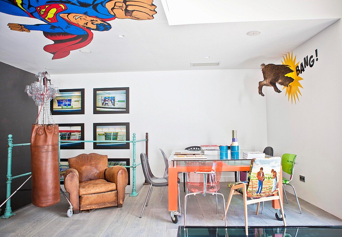 Various chairs around table with glass top and castors, vintage leather armchair and punching bag in interior with comic-book mural on ceiling and wall
