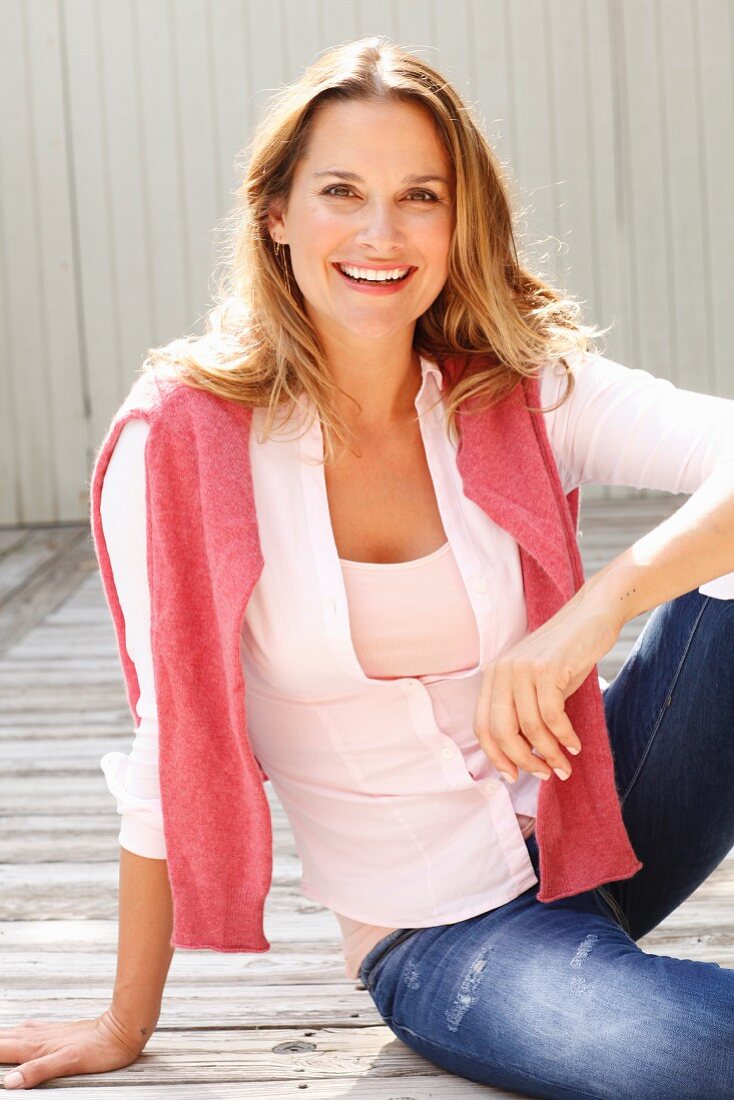 A blonde woman wearing a pink top, blouse and jeans with a pink jumper over her shoulders