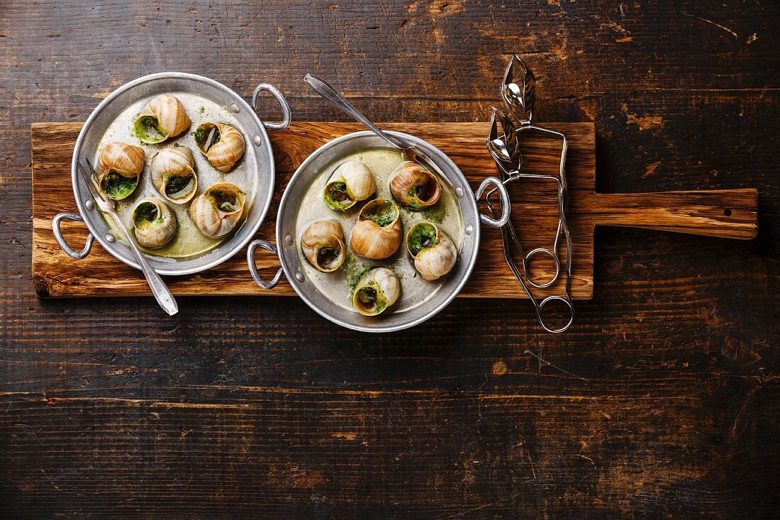 Escargots de Bourgogne (Weinbergschnecken mit Kräuterbutter) in Aluminiumpfanne auf rustikalem Holzbrett