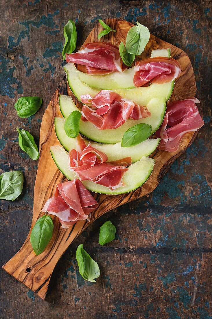 Sliced melon with ham and basil leaves, served on olive wood chopping board