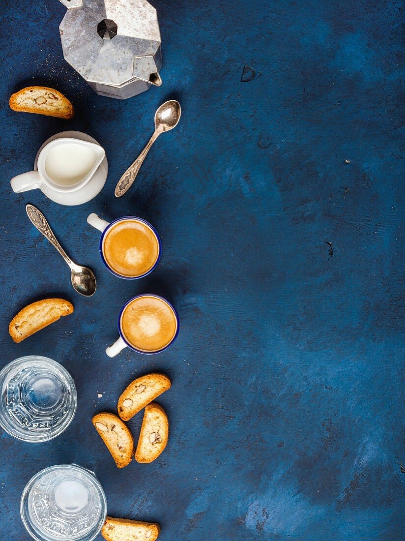Espressokanne, Cantucci, Milchkännchen, zwei Tassen Espresso und Wassergläser auf blauem Untergrund