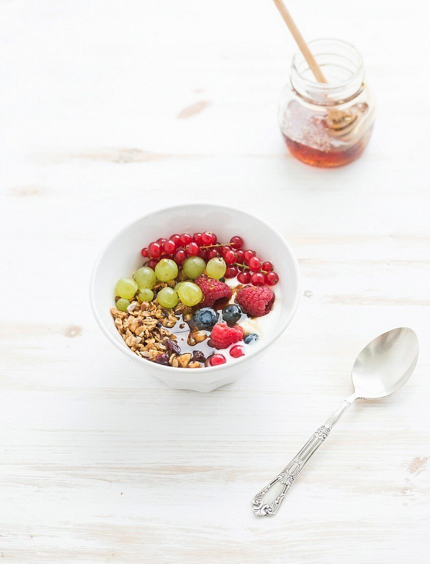 Oat granola crumble with fresh fruit, berres, honey in bowl over white painted wooden background