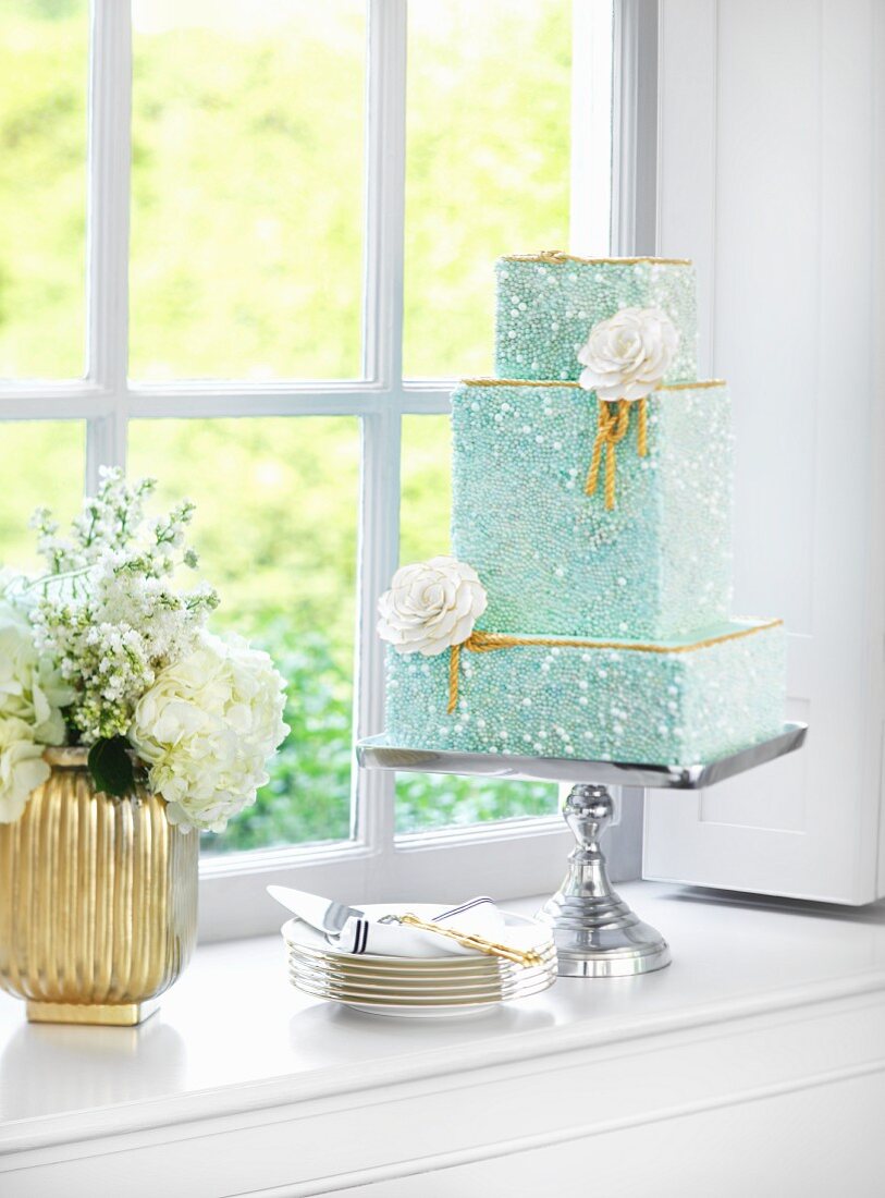 A turquoise three-tiered wedding cake on a silver cake stand in front of a window