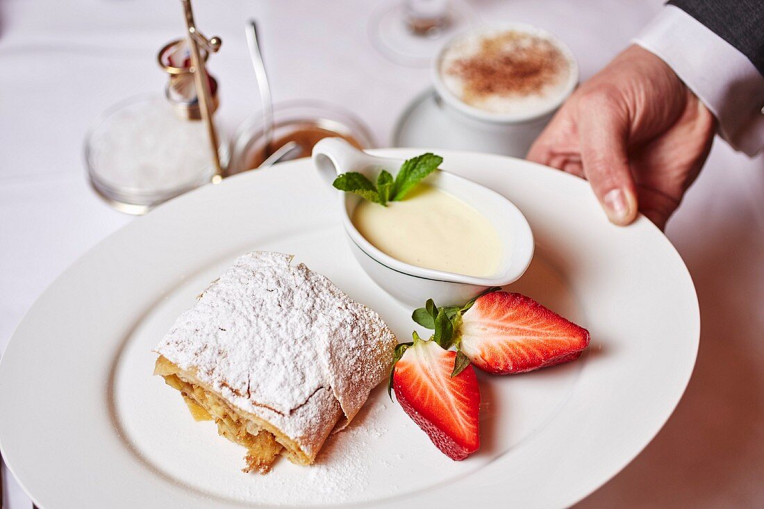 Kellner serviert Apfelstrudel mit Vanillesauce und Erdbeeren