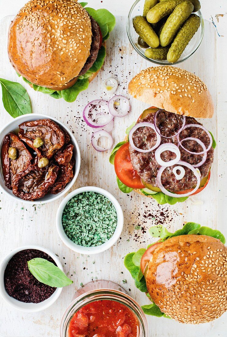 Selbst gemachte Rindfleisch-Burger mit Zwiebeln, daneben Gurken, Gewürze, getrocknete Tomaten und Tomatensauce in Schälchen