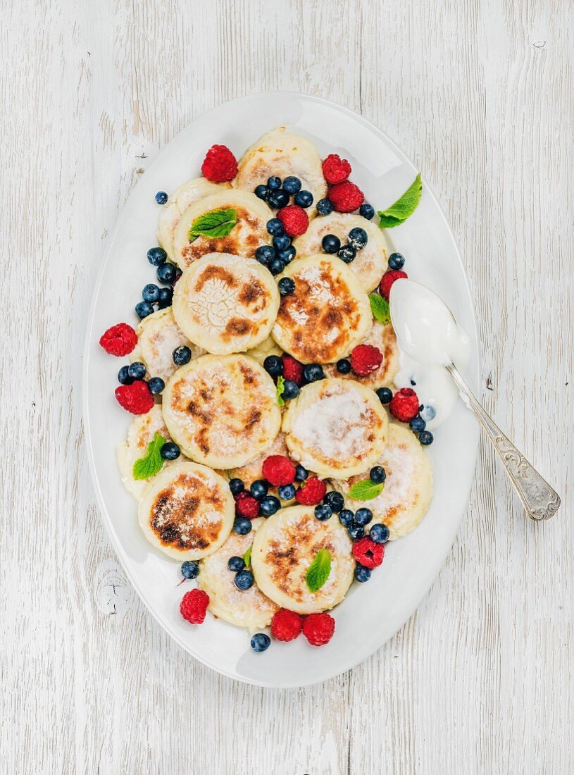 Syrniki (russische und ukrainische Pancakes aus Hüttenkäse), serviert mit frischen Waldbeeren und Sauerrahm