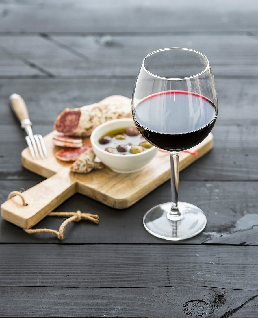 Glass of red wine, French sausage and olives on black wooden backdrop