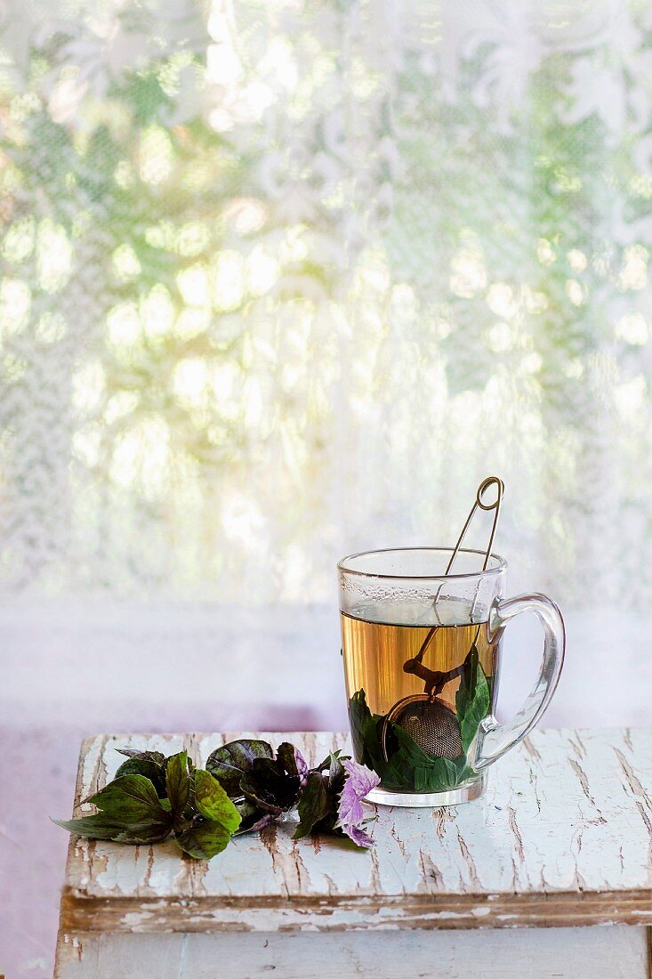 Kräutertee mit Teesieb in Glastasse, daneben frischer violetter Basilikum auf altem Holzhocker vor Fenster