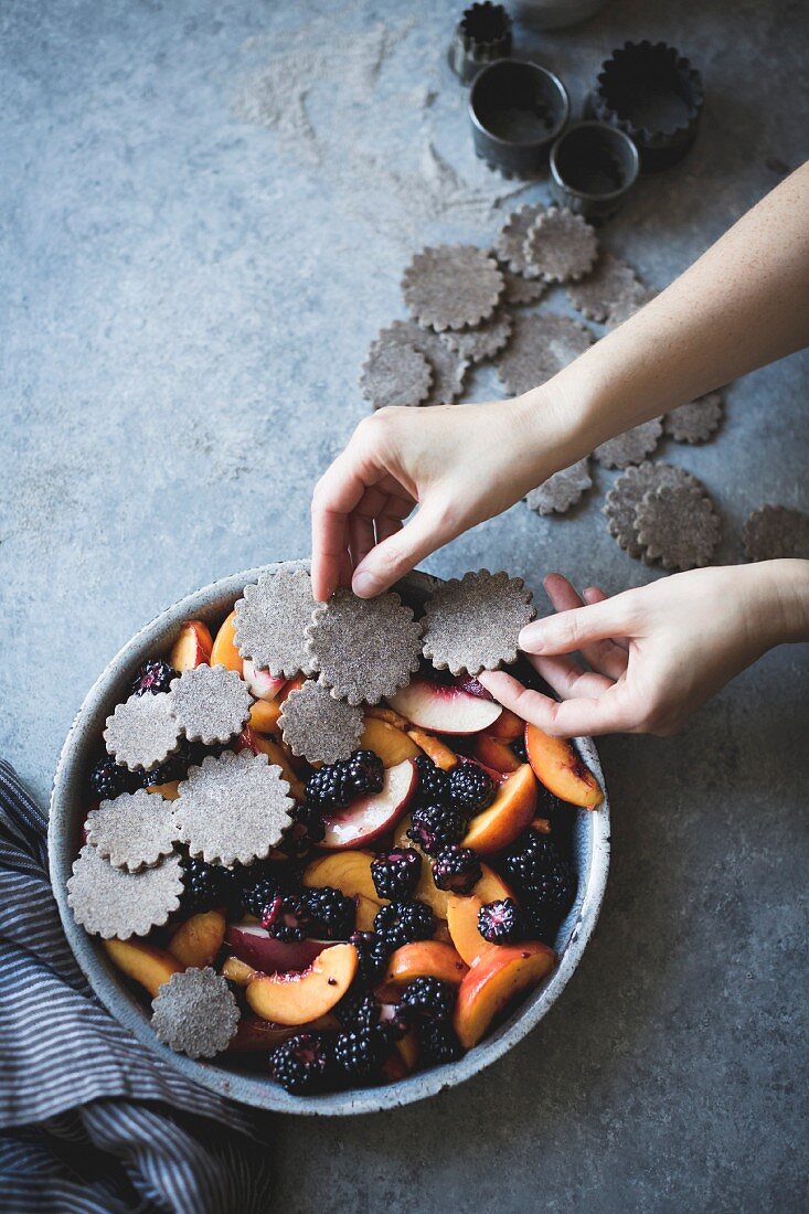 A hand placing dough shapes on a blackberry peach buckwheat pandowdy