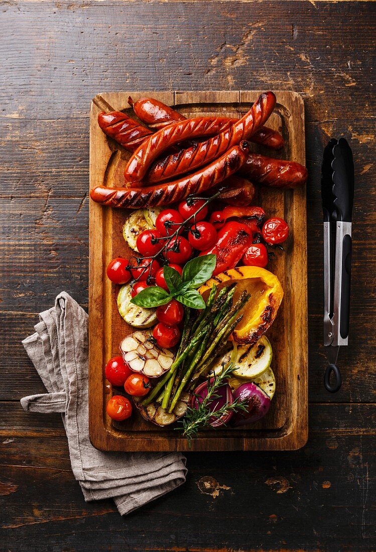 Grilled sausages and vegetables on cutting board on dark wooden background