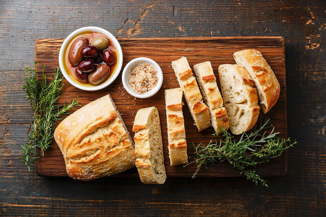 Fresh Ciabatta bread cut in slices on wooden cutting board with olives and herbs on rustic wooden background
