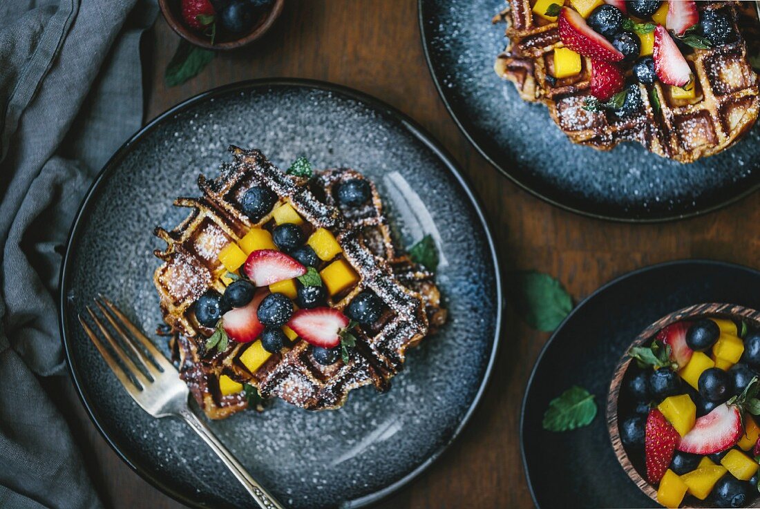 Two plates of brioche waffles are topped off with fruit and dusted with powdered sugar