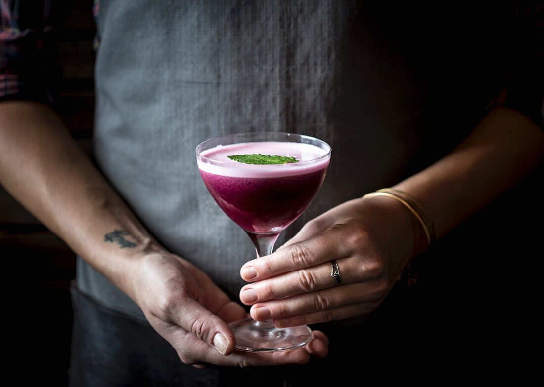 A woman holding a blueberry cocktail in a stemmed glass