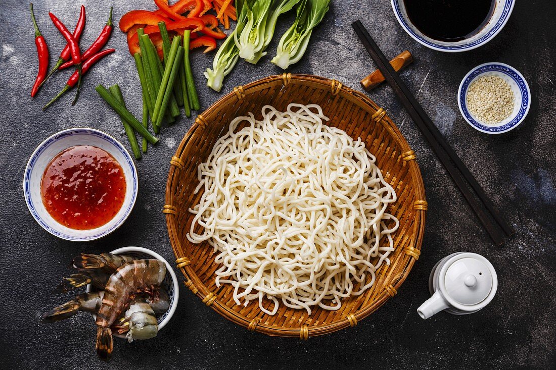 Raw Udon noodles in bamboo basket and Ingredients for cooking asian food with Tiger shrimps, greens, vegetables, spices on dark background