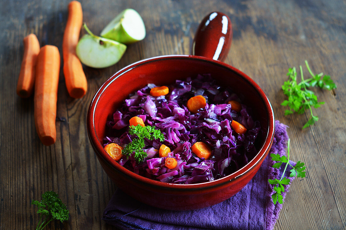 Red cabbage with carrots and apple in a ceramic pot with fresh ingredients in the background