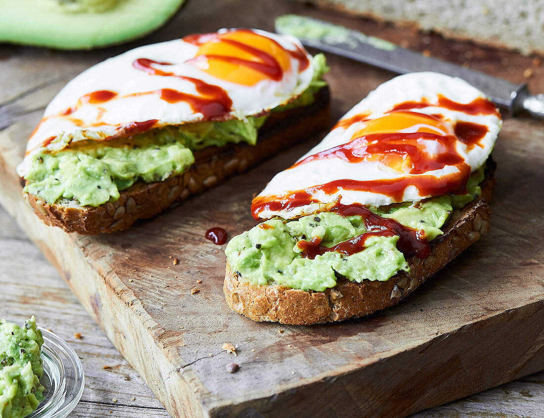 Two slices of bread topped with smashed avocado, a fried egg and chilli sauce
