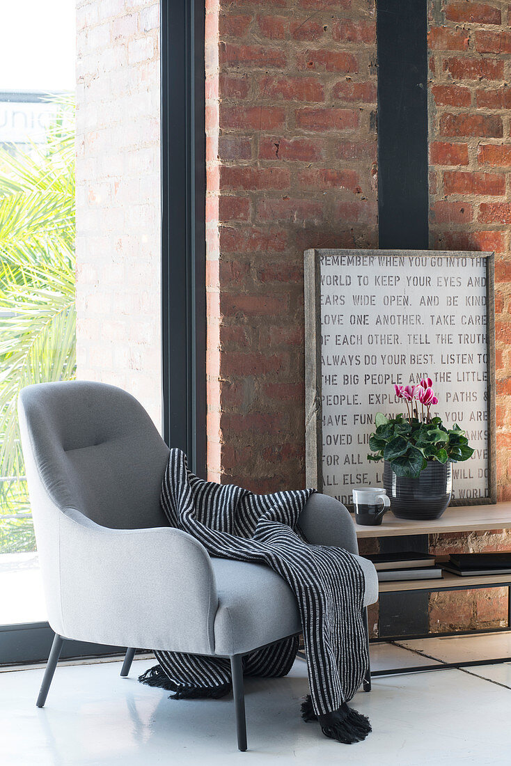 Blanket on grey armchair and potted cyclamen on low sideboard against brick wall