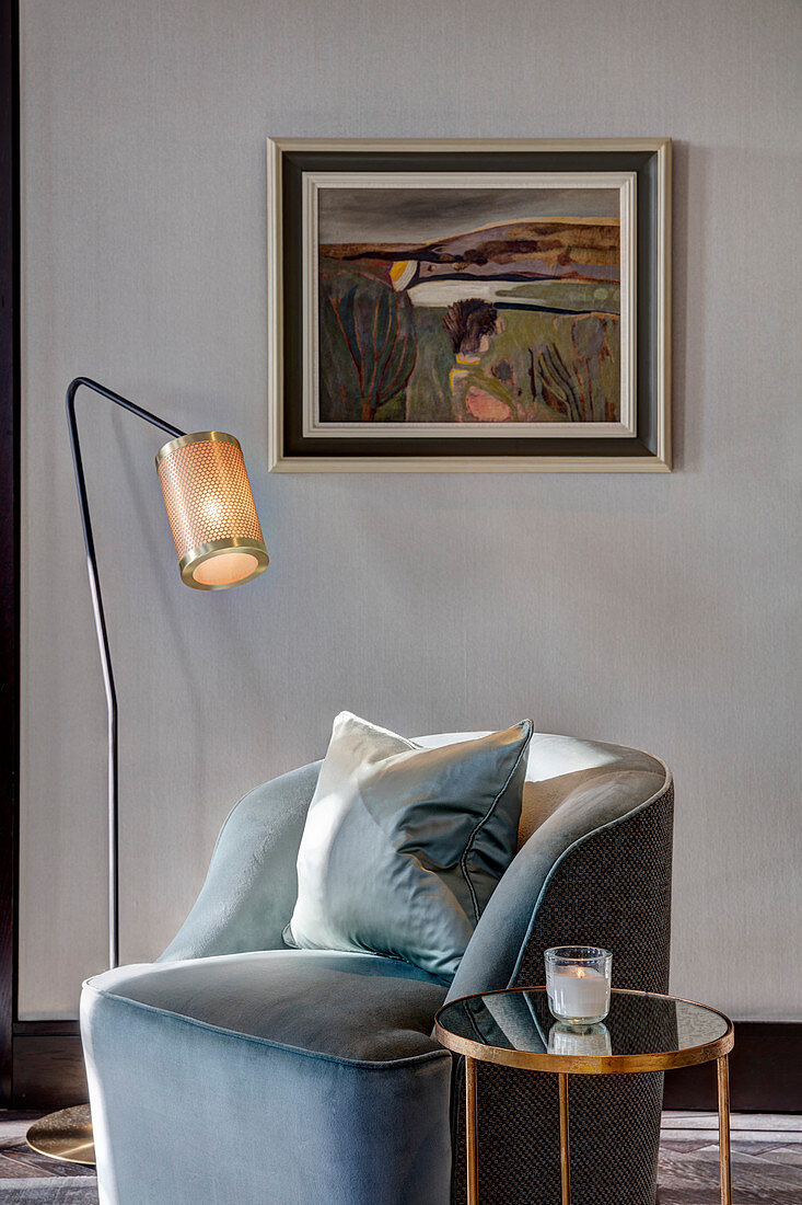 Armchair with scatter cushions between standard lamp and side table in front of painting on wall