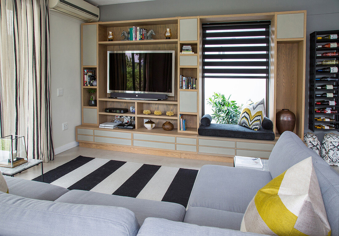 Regal mit Fernseher und integrierter Sitzbank vor Fenster, schwarz-weiss gestreifter Teppich und hellgraue Polstergarnitur im Wohnzimmer