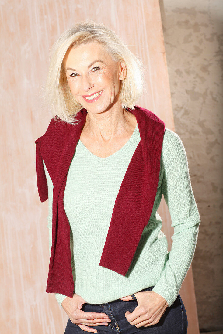 An older blonde woman wearing a light green knitted jumper with a dark red jumper over her shoulders