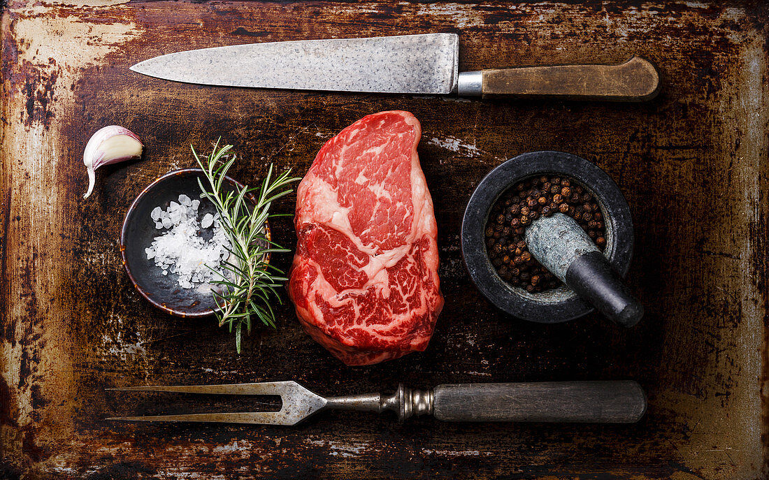 Rohes Rib Eye Steak mit Gewürzen und Fleischbesteck auf dunklem Untergrund