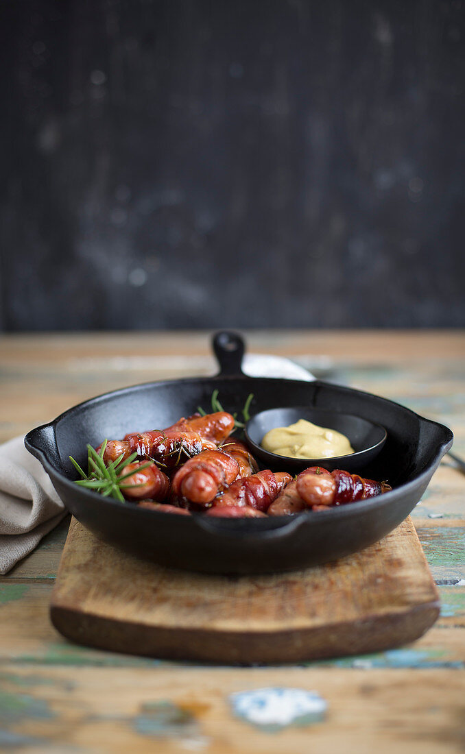 Chipolata sausages wrapped in bacon, served with mustard in a frying pan