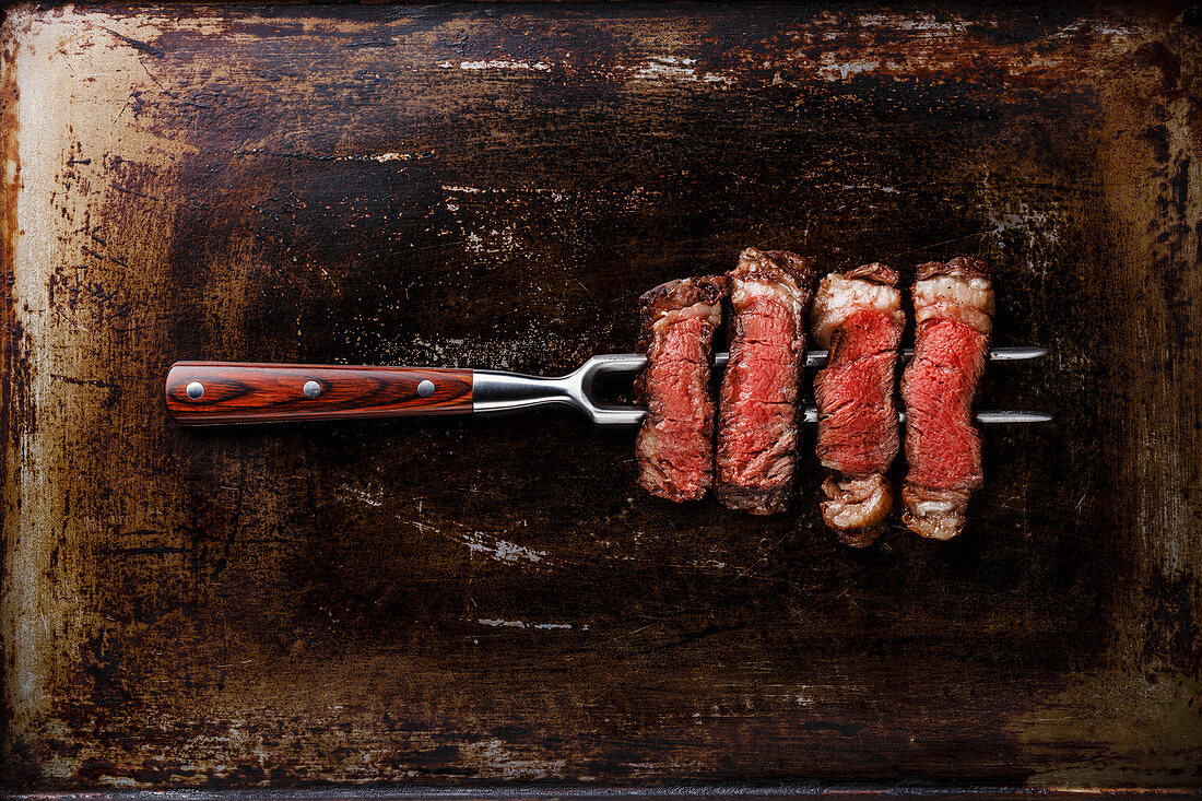 Gegrilltes Ribeye Steak in Scheiben auf Fleischgabel vor dunklem Metallhintergrund