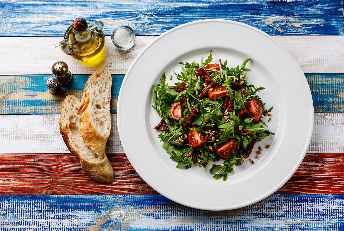 Salad with arugula, sun-dried tomatoes and sunflower seeds on white plate on wooden background